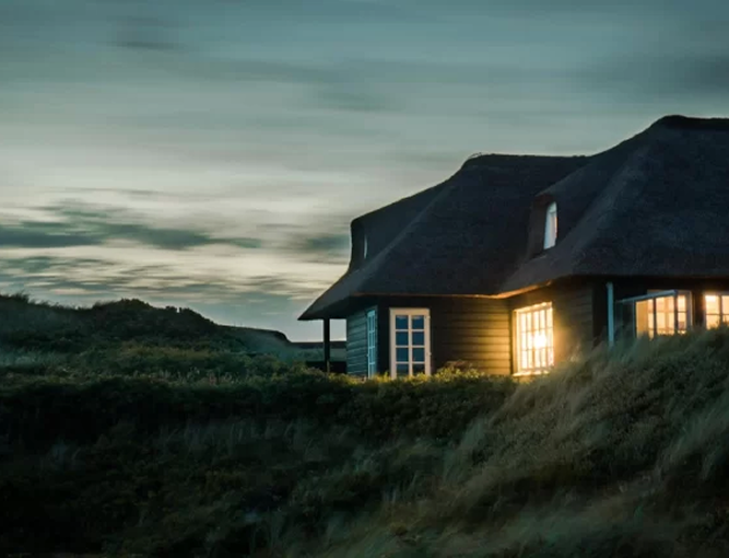 A cozy house with lights glowing inside sits among grassy hills at dusk, with a cloudy sky and fading daylight in the background.
